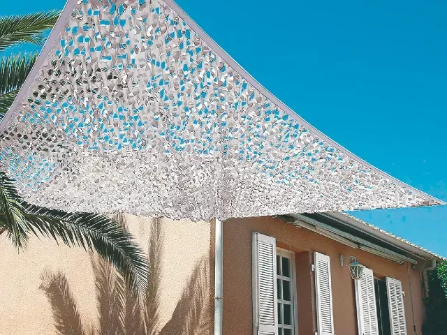 Voile d’ombrage triangulaire ajouré tendu au-dessus d’une terrasse méditerranéenne, en façade d’une maison aux volets blancs sous ciel bleu.