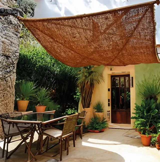 Terrasse ombragée avec voile en fibre naturelle, mobilier en bambou et végétation méditerranéenne devant une maison aux murs pastel.