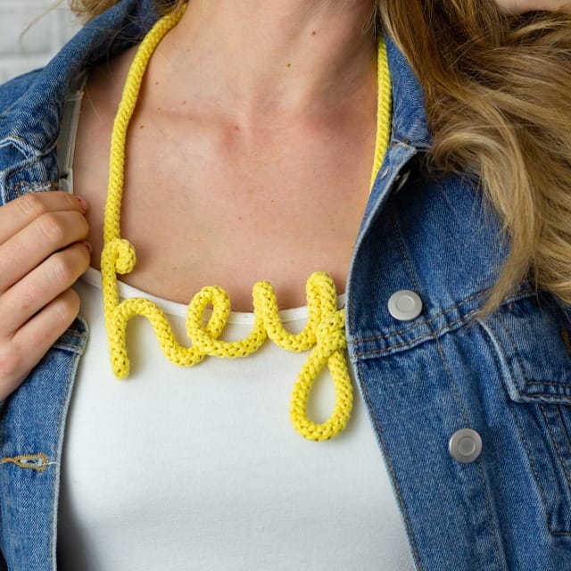 Collier en corde jaune formant le mot “hey”, porté avec un débardeur blanc et une veste en jean.