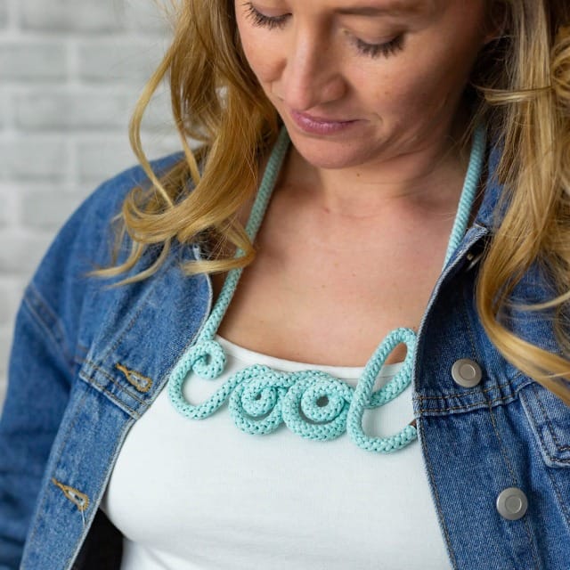 Collier en corde turquoise formant le mot “cool”, porté avec un débardeur blanc et une veste en jean.