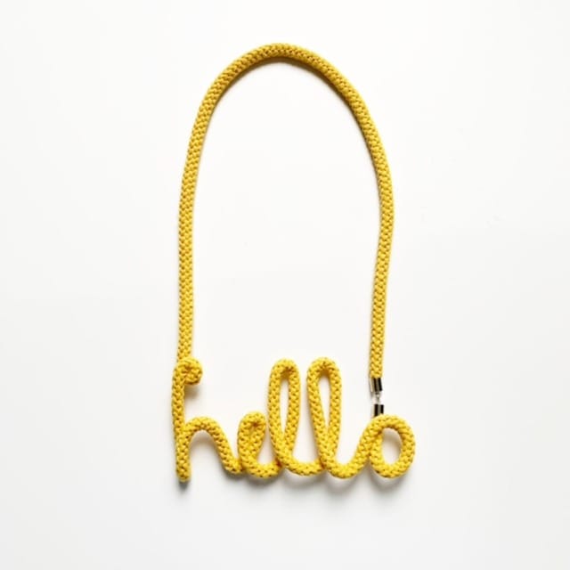 Collier en corde jaune formant le mot “hello”, sur fond blanc, bijou original au style graphique et ludique.
