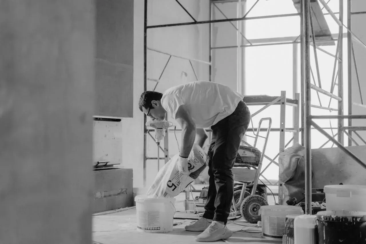 Artisan en plein geste versant un sac d’enduit dans un seau, sur un chantier intérieur avec échafaudages en arrière-plan