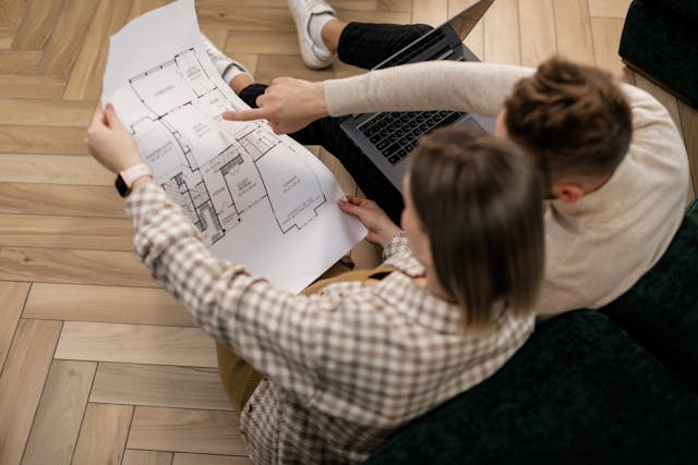 Couple assis par terre examinant un plan d’architecte étalé au sol, réflexion sur projet immobilier ou aménagement d’intérieur
