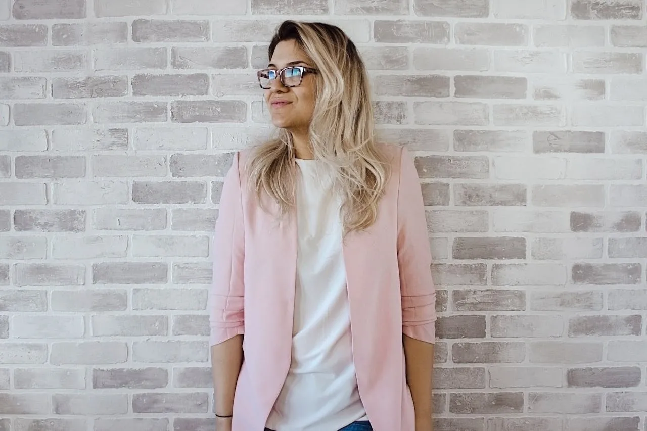Femme blonde portant un blazer rose poudré et un haut blanc, posant avec assurance devant un mur en briques claires