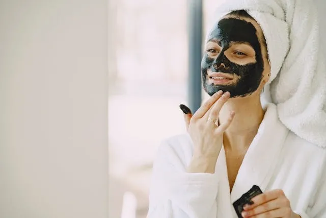 Femme appliquant un masque visage noir chez elle, rituel beauté cocooning en peignoir blanc.