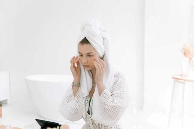 Femme en peignoir avec serviette sur la tête appliquant un soin visage, dans une salle de bain blanche à l’ambiance apaisée