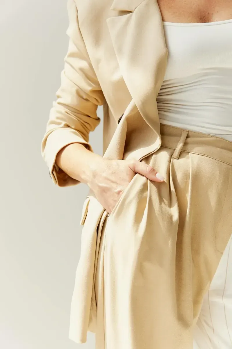 Femme en tailleur beige clair avec veste fluide et top blanc, debout avec la main glissée dans la poche du pantalon