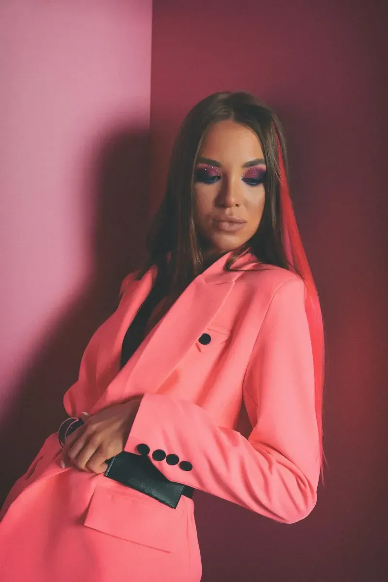Femme en tailleur rose fluo posant devant un mur bicolore, maquillage accentué et regard baissé, dans une composition visuelle percutante