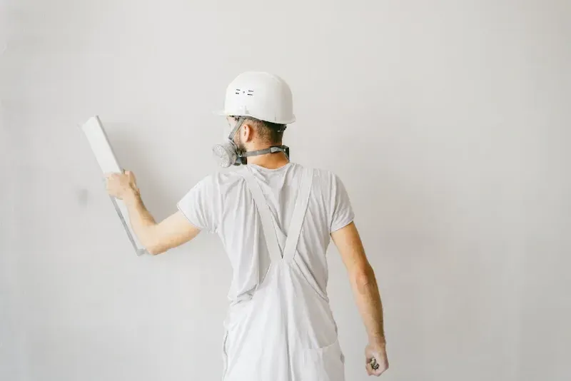 Ouvrier en tenue blanche appliquant soigneusement de l’enduit sur un mur, équipé d’un casque et d’un masque de protection