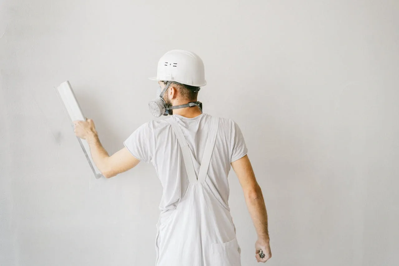Ouvrier en tenue blanche appliquant soigneusement de l’enduit sur un mur, équipé d’un casque et d’un masque de protection
