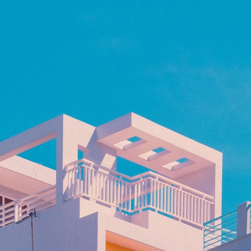 Terrasse minimaliste en béton blanc avec garde-corps géométrique, ouverte sur un ciel bleu intense et bordée de végétation méditerranéenne