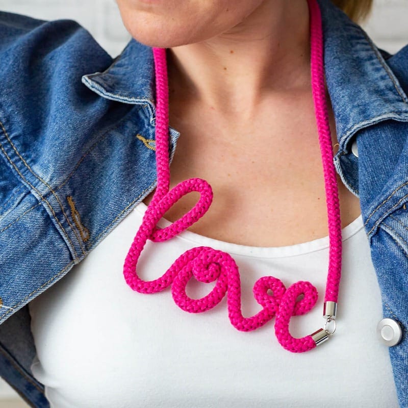 Collier en corde rose écrivant le mot “love”, porté avec un top blanc et une veste en jean.