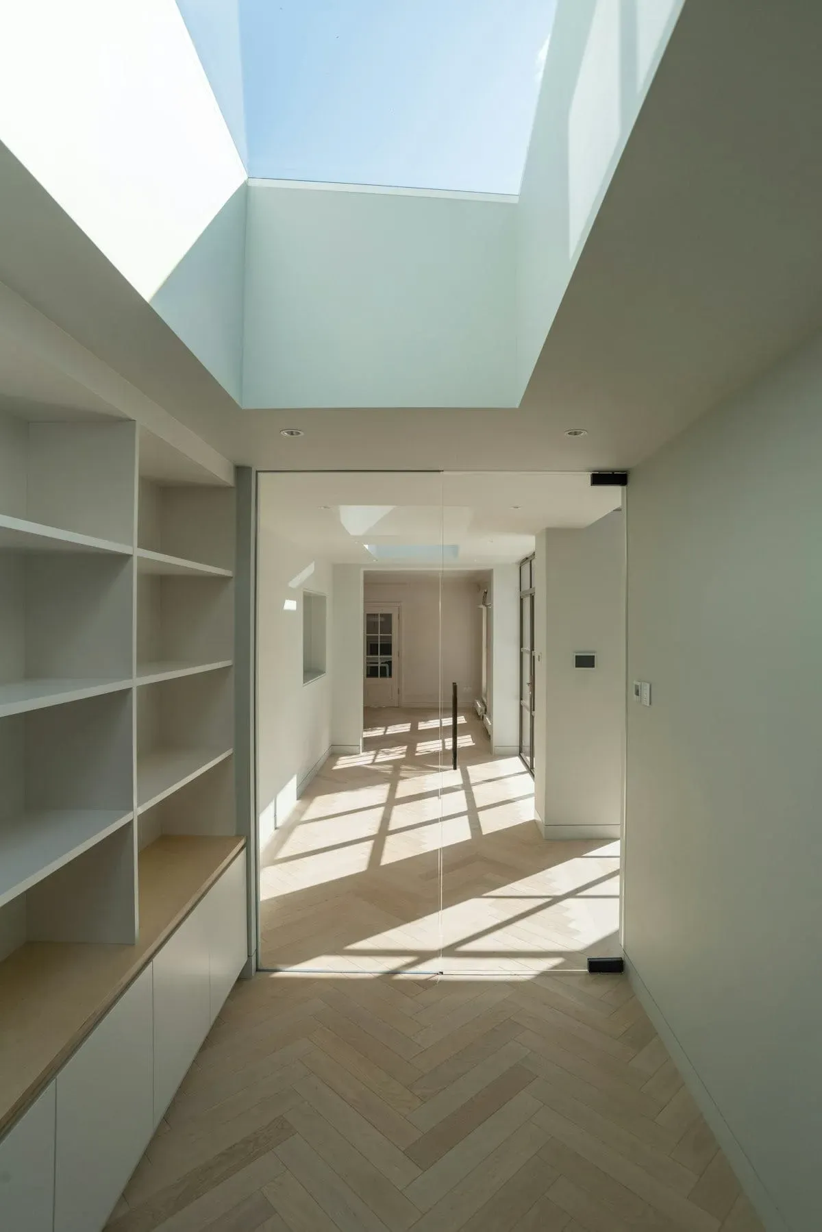 Couloir contemporain éclairé par une verrière zénithale, avec parquet en chevrons et aménagements intégrés blancs dans une atmosphère lumineuse