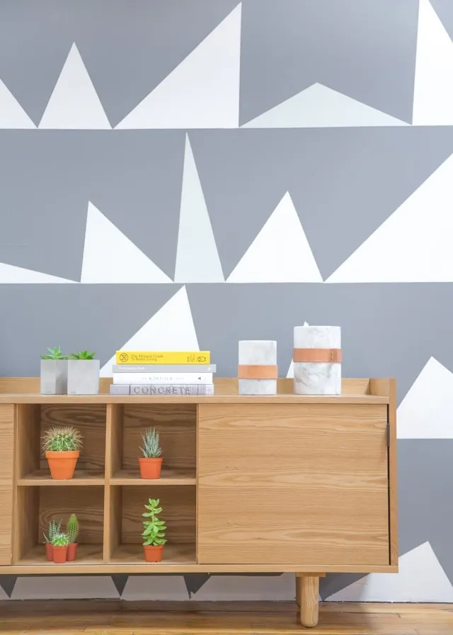 Enfilade en bois clair contre un mur graphique gris et blanc, décorée de petits cactus en pot, livres et objets minimalistes
