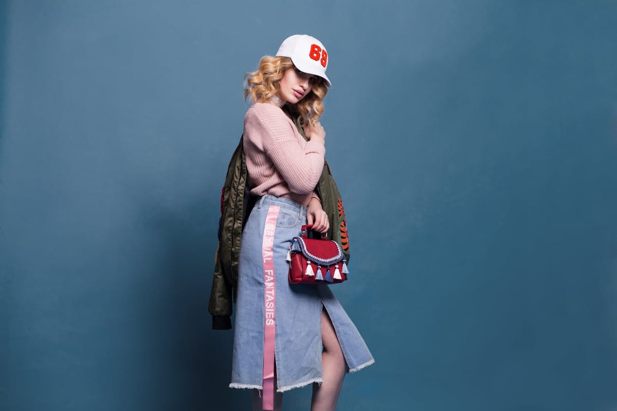 Femme blonde en jupe en jean et pull rose poudré, coiffée d’une casquette blanche et vêtue d’une veste kaki, posant sur fond bleu uni