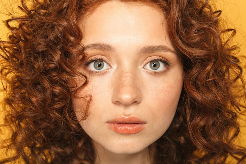 Portrait d’une jeune femme rousse aux cheveux bouclés, peau claire et taches de rousseur sur fond jaune doux