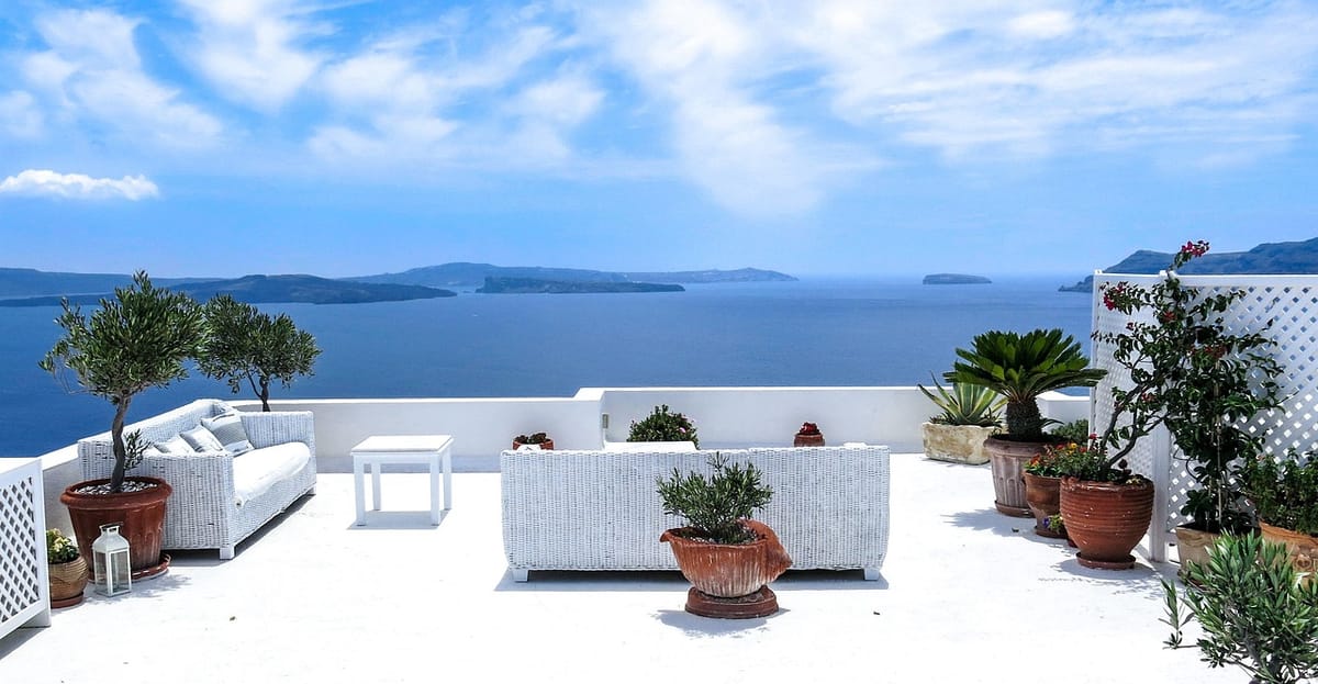 Terrasse baignée de soleil avec mobilier en rotin blanc, plantes en pot et vue panoramique sur la mer sous un ciel lumineux