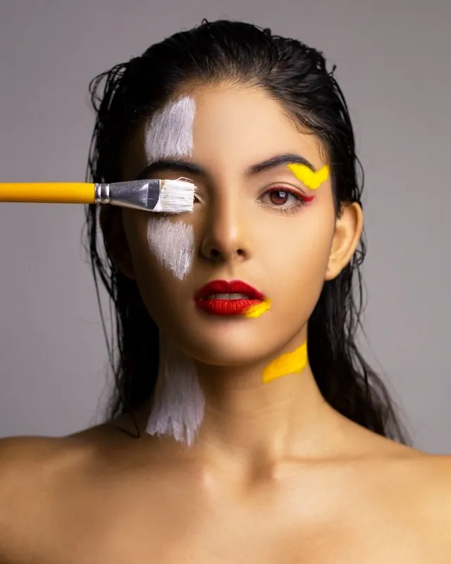 Femme au visage peint de blanc et jaune, maquillage artistique appliqué au pinceau, regard intense