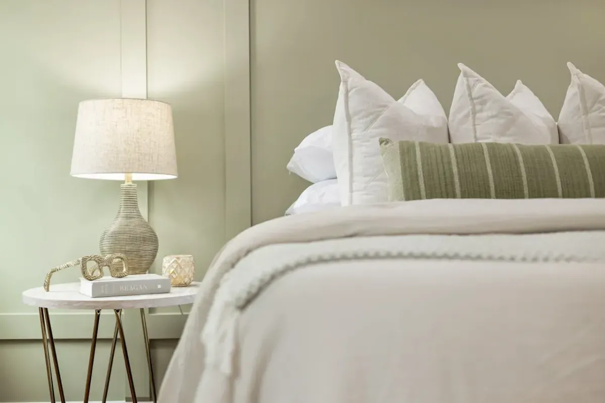 Chambre lumineuse avec lampe en rotin, linge de lit blanc et coussin vert rayé, décoration naturelle.
