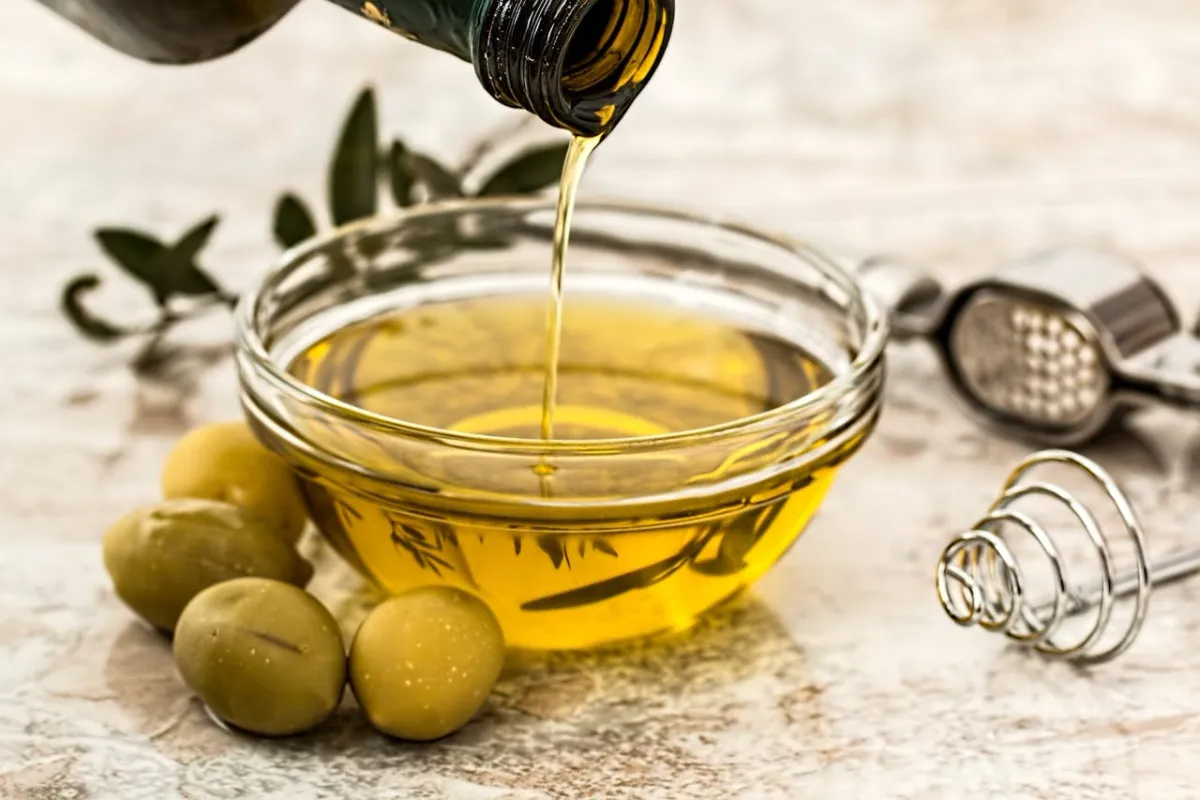 Huile d’olive dorée versée dans un bol en verre avec olives vertes sur une table en marbre