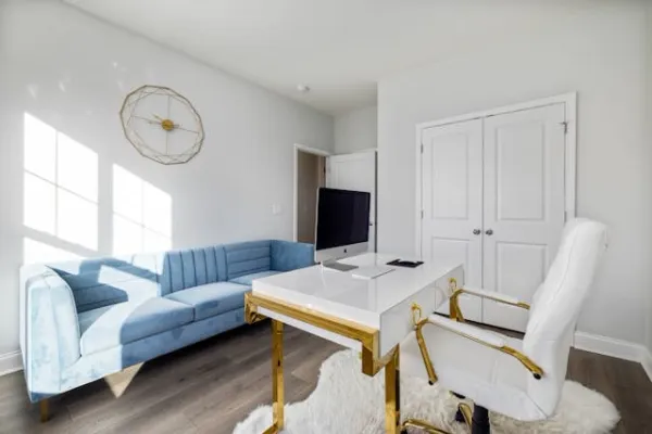 Bureau élégant avec mobilier blanc et doré, fauteuil capitonné crème, canapé bleu pastel et déco murale dorée géométrique sous lumière naturelle