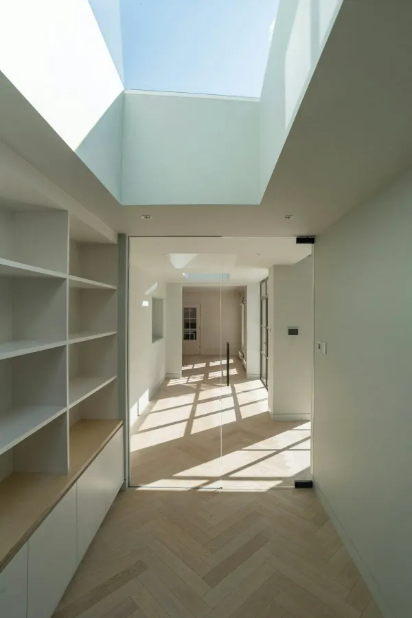 Couloir contemporain éclairé par une verrière zénithale, avec parquet en chevrons et aménagements intégrés blancs dans une atmosphère lumineuse