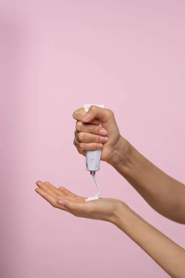 Crème fluide versée dans la paume d’une main depuis un tube blanc, geste de soin minimaliste sur fond rose doux