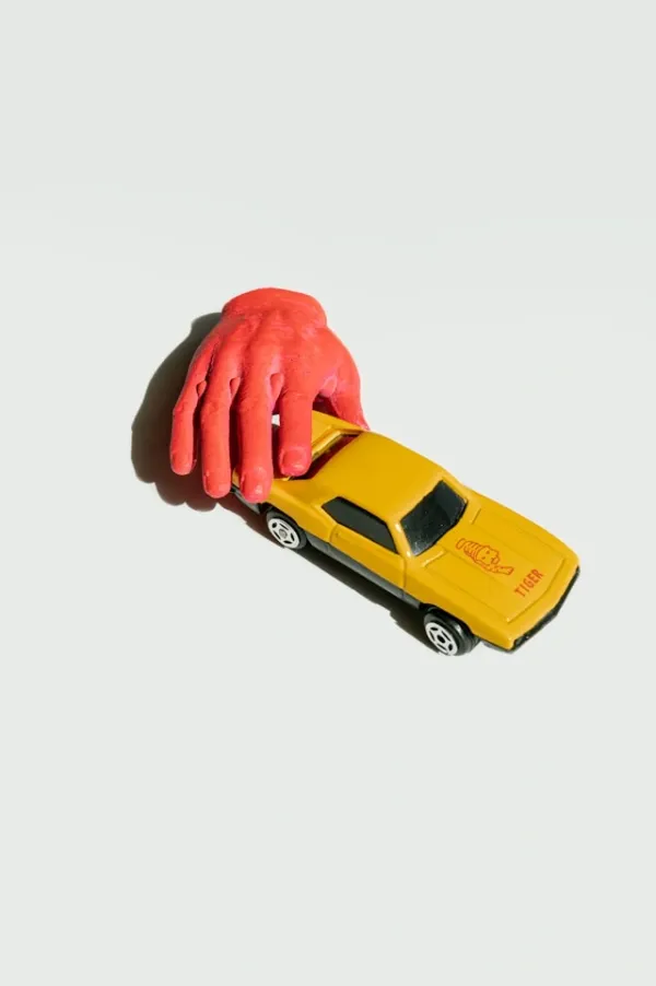 Main rouge en plastique posée sur une voiture miniature jaune, composition pop en lumière directe