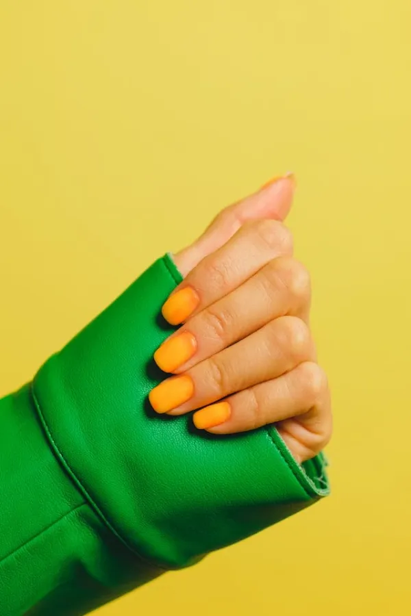 Main avec ongles vernis orange vif posée sur fond jaune, contraste mode avec veste verte et pose créative colorée