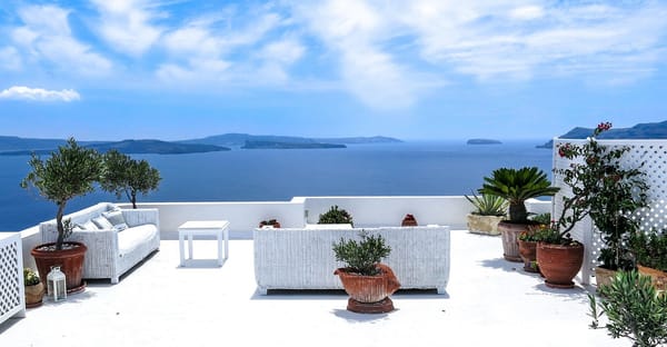 Terrasse baignée de soleil avec mobilier en rotin blanc, plantes en pot et vue panoramique sur la mer sous un ciel lumineux