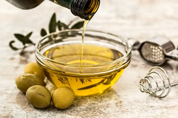 Huile d’olive dorée versée dans un bol en verre avec olives vertes sur une table en marbre