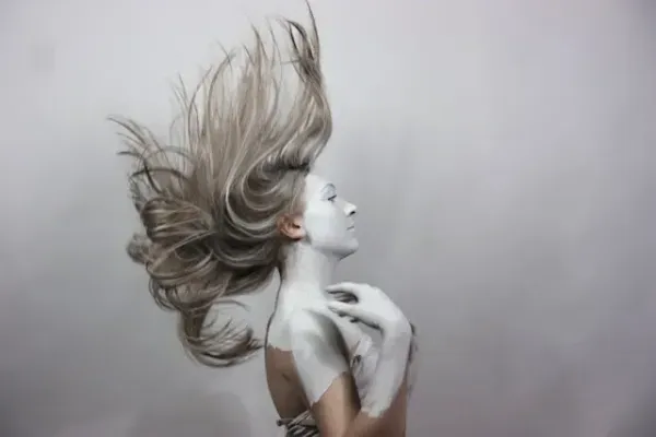 Femme au visage partiellement peint en blanc, cheveux projetés en l'air dans une pose artistique expressive.