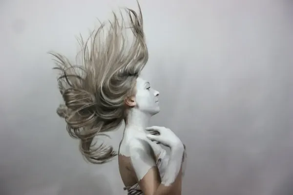 Femme au visage partiellement peint en blanc, cheveux projetés en l'air dans une pose artistique expressive.