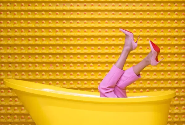 Jambes féminines en talons hauts roses sortant d’une baignoire jaune sur fond de canards en plastique