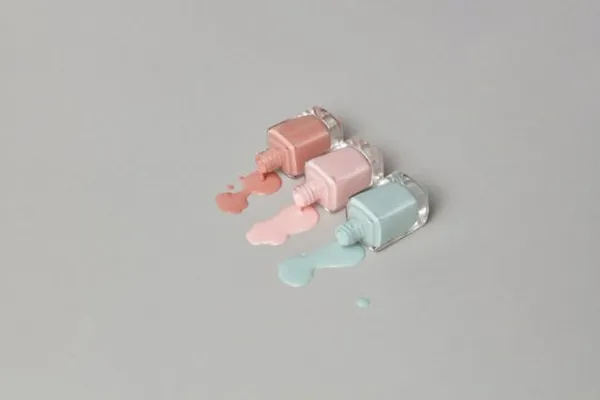 Trois flacons de vernis à ongles pastel renversés sur fond gris, teintes rose, nude et bleu clair